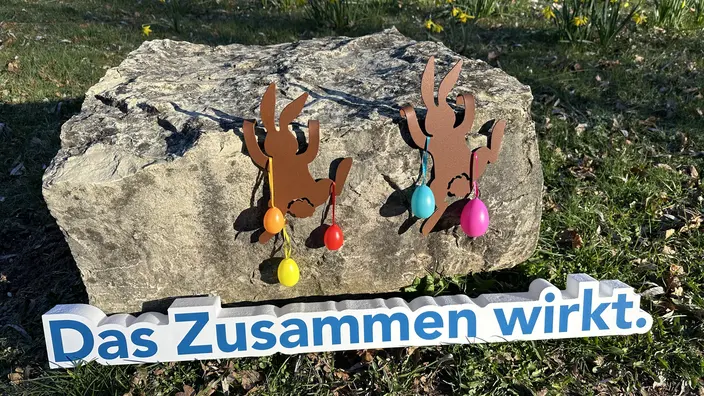 Der Slogan "Das Zusammen wirkt" vor einem österlich geschmückten Stein
